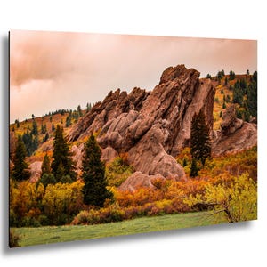 May include: A landscape photograph featuring a large rock formation in shades of brown and tan, set against a cloudy, peach-colored sky. The foreground shows green grass and colorful autumn foliage.