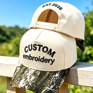 Könnte beinhalten: Eine cremefarbene Baseballkappe mit einem grün-weißen Tarnmuster auf dem Schirm. Die Vorderseite zeigt den schwarzen Sticktext CUSTOM embroidery, während der hintere Riemenbereich den schwarzen Sticktext EST. 2026 aufweist.