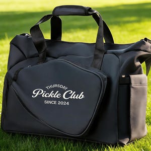 Könnte beinhalten: Schwarze Tragetasche mit abnehmbarer Tasche. Die Tasche und der Beutel tragen den Text "Thursday Pickle Club SINCE 2024" in weißer Schrift. Die Tasche hat einen oberen Griff und Schultergurte, auf grünem Gras.