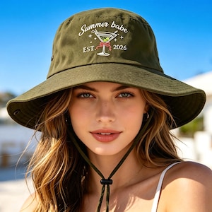 May include: An olive green bucket hat featuring an embroidered graphic of a martini glass with olives and a red bow. The text reads Summer babe EST. 2026. The hat includes a matching olive green chin strap with an adjustable black toggle fastener.