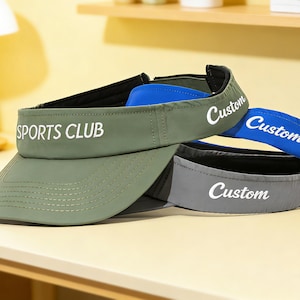 Könnte beinhalten: Drei Sonnenblenden in Olivgrün, Blau und Grau werden gezeigt. Auf der olivgrünen Sonnenblende steht "SPORTS CLUB". Auf den anderen beiden Sonnenblenden steht "Custom". Die Sonnenblenden sind aus leichtem Material gefertigt.