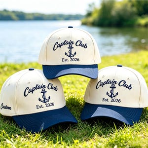 Puede incluir: Tres gorras de béisbol con corona color crema y visera azul marino. Cada gorra presenta un bordado azul oscuro que dice Captain Club con un icono de ancla y el texto Est. 2026. Una gorra también muestra la palabra Custom bordada en el lateral.