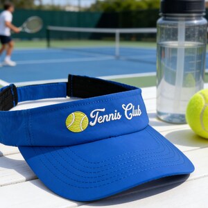 May include: A blue tennis visor with white embroidered text reading "Tennis Club" and a yellow tennis ball emblem. A clear water bottle and a yellow tennis ball are also present.