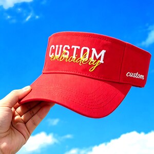 Può includere: Una visiera rosso brillante con la parola CUSTOM in grandi lettere maiuscole bianche e la parola embroidery scritta in corsivo giallo sottostante. Il lato della visiera mostra anche la parola custom in piccole lettere minuscole bianche.