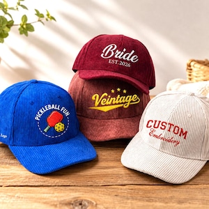 May include: A collection of four corduroy baseball caps in blue, maroon, and cream. The blue cap features a pickleball graphic and text. The maroon caps display white 'Bride EST.2026' and yellow 'Veintage' text. The cream cap features 'CUSTOM Embroidery' text.