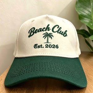 Puede incluir: Gorra de béisbol blanca y verde con la inscripción bordada "Beach Club" en verde, un gráfico de palmera y el texto "Est. 2026". La gorra tiene una visera verde.