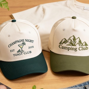 May include: Two baseball caps with curved peaks. One is cream-coloured with a dark green peak and the text "Champagne Night Toast Club" with a cocktail graphic. The other is cream-coloured with a green peak and the text "Camping Club" with a mountain graphic.
