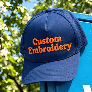 以下が含まれることがあります： ネイビーブルーの野球帽で、盛り上がったオレンジ色の文字で「Custom Embroidery」と刺繍されています。帽子は構造化されたクラウン、上部のボタン、通気用のパンチング加工されたサイドパネル、そしてカーブしたつばが特徴です。
