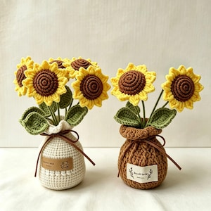 May include: Two crocheted sunflower arrangements in textured pots. One pot is cream with a brown leather-like tag featuring a bear icon, the other is brown with a white fabric tag showing a leaf design. Both have brown ties.