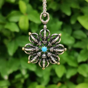 Pode incluir: Colar com pingente prateado com um design detalhado e ornamental. O pingente tem a forma de uma flor estilizada com uma pedra central de cor turquesa. As pontas das pétalas da flor têm detalhes dourados. A corrente do colar também é prateada.