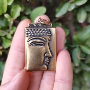 May include: A rectangular, bronze-colored pendant featuring a detailed relief of Buddha's profile. The pendant has a loop at the top for a chain and is held in a hand against a blurred green background. The Buddha's face is the focal point.