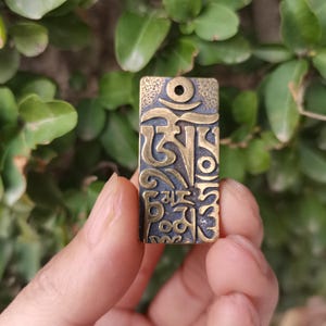 Puede incluir: Colgante rectangular de latón con escritura tibetana grabada. El colgante tiene un acabado oscuro y envejecido con letras en relieve de color dorado y un pequeño agujero en la parte superior para una cadena o cordón. El fondo es verde borroso.