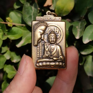 May include: A rectangular bronze pendant featuring a detailed relief of Buddha. The pendant has a loop at the top for a chain. The design includes a Buddha figure and Chinese characters. The background is a natural green.