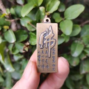 May include: A rectangular brass pendant with a loop at the top. The pendant features embossed Chinese characters and figures. The background is a blurred view of green leaves.