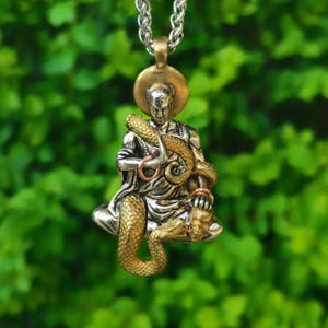 Può includere: Un ciondolo color argento raffigurante una figura seduta con aureola, drappeggiata in vesti e intrecciata con un serpente color oro. La figura tiene un piccolo teschio in una mano ed è appesa a una collana a catena in metallo argentato.