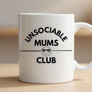 Puede incluir: Taza de cerámica blanca con el texto negro "UNSOCIABLE MUMS CLUB", con una cara sonriente con gafas de sol. La taza tiene un asa curva y está colocada sobre una superficie de madera clara.