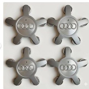 May include: Four gray Audi wheel center caps arranged on a white surface. Each cap features the Audi logo in the center and five star-shaped extensions with mesh detailing. The caps are metallic and have a matte finish.