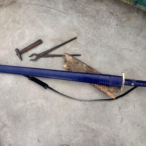 May include: A long sword encased in a deep blue leather scabbard with a metallic tip and pommel. The sword features a gold-colored crossguard and a black leather carrying strap. Nearby are a small metal hammer and a pair of metal tongs.