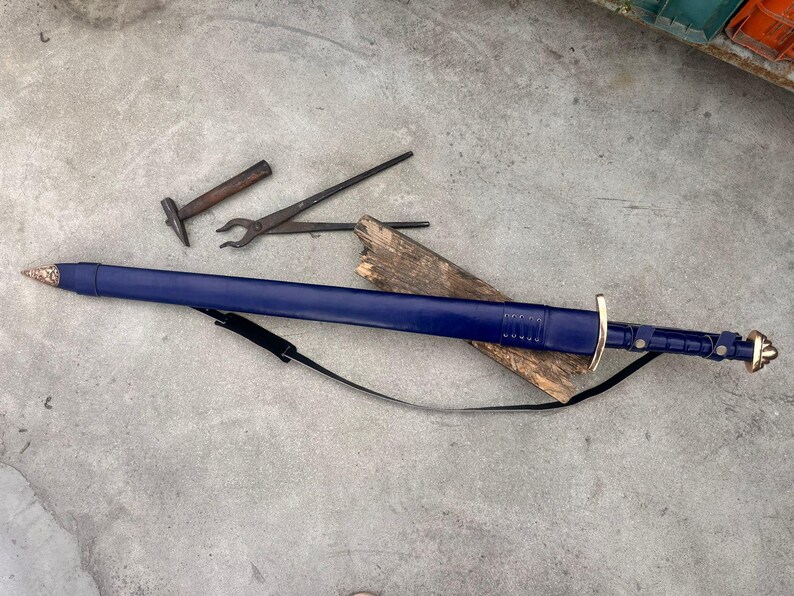 May include: A long sword encased in a deep blue leather scabbard with a metallic tip and pommel. The sword features a gold-colored crossguard and a black leather carrying strap. Nearby are a small metal hammer and a pair of metal tongs.