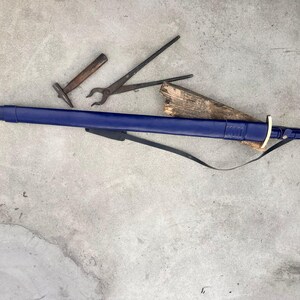 May include: A long sword encased in a deep blue leather scabbard with a metallic tip and pommel, featuring a gold-colored crossguard and a black leather strap. Nearby are a small metal hammer and a pair of black iron blacksmith tongs.