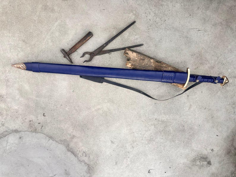 May include: A long sword encased in a deep blue leather scabbard with a metallic tip and pommel, featuring a gold-colored crossguard and a black leather strap. Nearby are a small metal hammer and a pair of black iron blacksmith tongs.