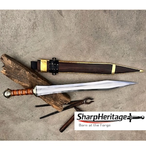 May include: A double-edged sword with a polished steel blade and a ribbed wooden handle featuring a brass pommel. The accompanying scabbard is made of dark wood with brass accents and a black leather strap. The text SharpHeritage Born at the Forge.