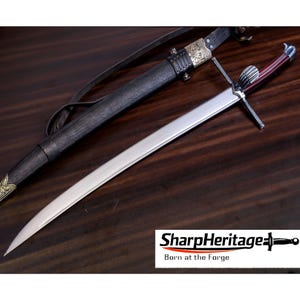 Pode incluir: Uma espada de aço com cabo castanho-avermelhado escuro, guarda prateada e pomo canelado. A espada é acompanhada por uma bainha de couro preto com ornamentos metálicos dourados e uma correia de couro. O texto diz SharpHeritage.