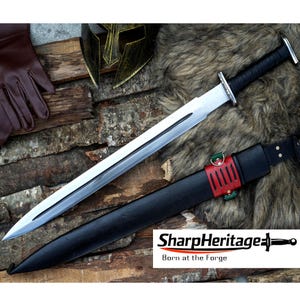 May include: A long, silver sword with a black handle and a black leather sheath. The sword rests on a wooden surface with a brown leather glove, a gold helmet, and a fur rug. The text "SharpHeritage" is visible.