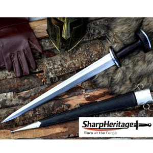 May include: A Viking sword with a polished steel blade and a dark wooden handle, resting on a wooden surface. The sword is accompanied by a black leather sheath with silver accents. The image also includes a brown leather glove and a gold helmet. The text "SharpHeritage" is visible.