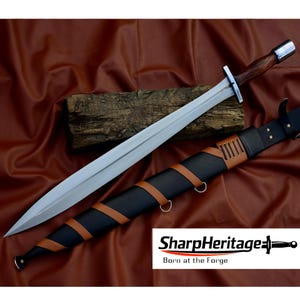 May include: A long, silver sword with a wooden handle and a metal crossguard. It rests on a brown surface. The sword is sheathed in a black leather scabbard with brown leather accents. The text "SharpHeritage" and "Born at the Forge" are visible.