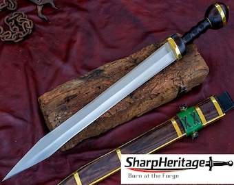 Puede incluir: Una espada de doble filo con hoja de acero pulido, empuñadura de madera oscura y pomo redondeado con detalles en latón. La vaina a juego presenta madera oscura, bandas de latón y una sección de cuero verde. El texto dice SharpHeritage.