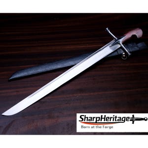 Puede incluir: Una espada recta de acero con hoja pulida, guarda de plata y mango de madera. La espada va acompañada de una vaina negra texturizada. La imagen incluye el texto SharpHeritage Born at the Forge con un icono de espada.
