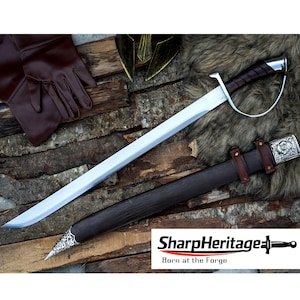 Puede incluir: Una espada plateada y marrón con una hoja curva, empuñadura forrada en cuero y una guarda decorativa plateada. La espada está en una vaina marrón oscuro con detalles plateados. También se muestran guantes marrones y un casco dorado.
