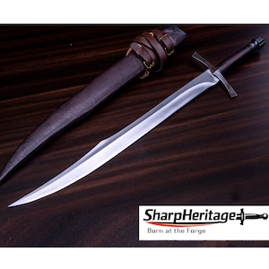 Puede incluir: Una espada de acero con hoja de doble filo, guarda recta y empuñadura oscura, acompañada de una vaina de cuero marrón con correas integradas y hebillas de latón. La imagen incluye el texto SharpHeritage Born at the Forge.