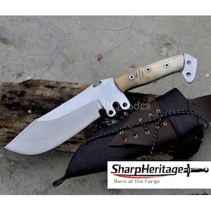 May include: A large, stainless steel knife with a bone handle and a black leather sheath. The knife has a wide blade and a curved edge. The sheath is secured with a laced cord. The text "SharpHeritage" and "Born at the Forge" are visible.