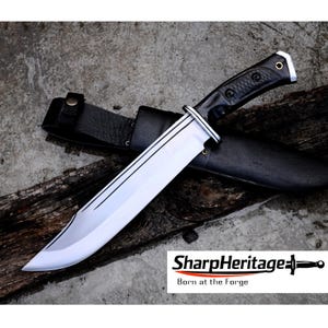 May include: A large, silver-bladed knife with a black handle and a leather sheath. The knife has a polished blade with a sharp edge and is resting on a piece of wood. The text "SharpHeritage Born at the Forge" is visible.