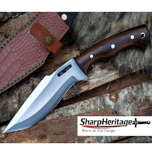 May include: A hunting knife with a polished steel blade and a dark wood handle. The knife has a full tang construction and is accompanied by a brown leather sheath with green stitching. The text "SharpHeritage Born at the Forge" is visible.