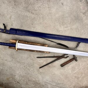 May include: A steel sword with a blue grip and gold-colored pommel and crossguard, resting on a wooden block. Beside it lies a matching blue leather scabbard with a metal tip and black straps, along with a pair of metal tongs and a small hammer.