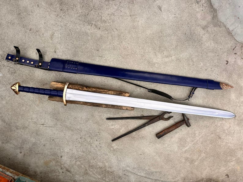 May include: A steel sword with a blue grip and gold-colored pommel and crossguard, resting on a wooden block. Beside it lies a matching blue leather scabbard with a metal tip and black straps, along with a pair of metal tongs and a small hammer.