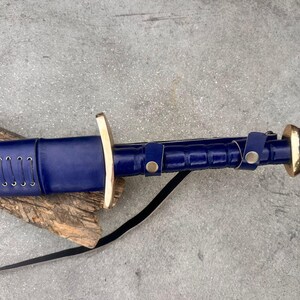 May include: A sword with a deep blue leather-wrapped scabbard and handle, featuring a polished brass crossguard and pommel. The scabbard is detailed with silver-toned lacing, while the handle includes two blue leather straps secured with brass snaps.