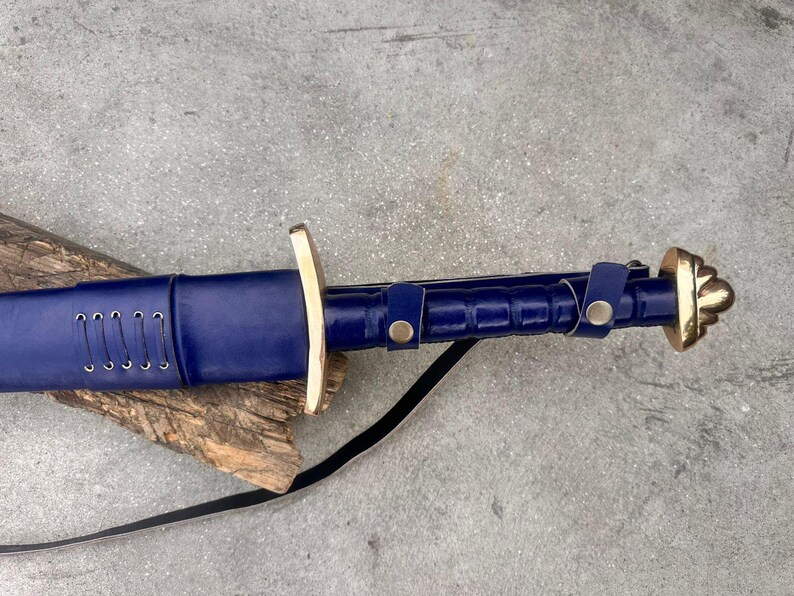 May include: A sword with a deep blue leather-wrapped scabbard and handle, featuring a polished brass crossguard and pommel. The scabbard is detailed with silver-toned lacing, while the handle includes two blue leather straps secured with brass snaps.