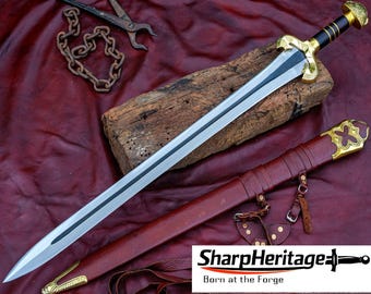 Puede incluir: Una espada de acero de doble filo con un canal central, que presenta una empuñadura negra con anillos dorados y una guarda dorada. La espada se acompaña de una vaina de cuero marrón con herrajes de metal dorado y correas de cuero.