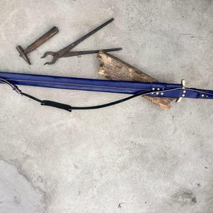 May include: A long, dark blue leather sword scabbard with a metallic tip and brass-colored rivets, featuring an attached black leather shoulder strap. Nearby are a small metal hammer and a pair of long-handled metal blacksmith tongs.