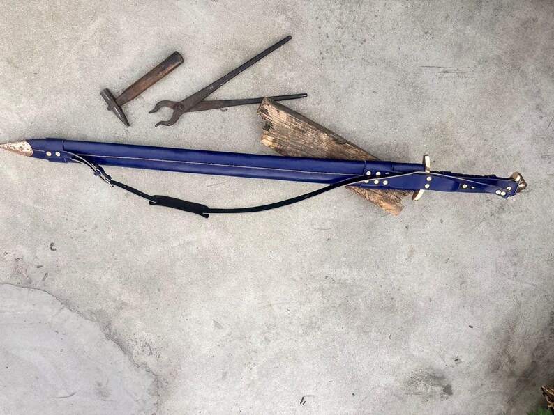 May include: A long, dark blue leather sword scabbard with a metallic tip and brass-colored rivets, featuring an attached black leather shoulder strap. Nearby are a small metal hammer and a pair of long-handled metal blacksmith tongs.