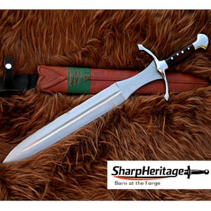 Könnte beinhalten: Ein silbernes und schwarzes Schwert mit braunem Griff und Lederscheide. Das Schwert hat eine zweischneidige Klinge und einen Parier. Die Scheide ist braun mit grünen Akzenten. Der Text "SharpHeritage Born at the Forge" ist enthalten.