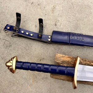 May include: A sword with a blue grip and a gold-colored pommel and crossguard, accompanied by a matching blue scabbard featuring black leather straps with gold-colored metal rivets and a laced detail along the upper section of the scabbard.