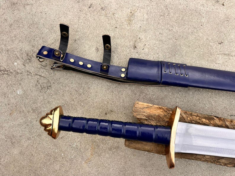 May include: A sword with a blue grip and a gold-colored pommel and crossguard, accompanied by a matching blue scabbard featuring black leather straps with gold-colored metal rivets and a laced detail along the upper section of the scabbard.