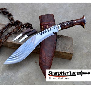 Puede incluir: Un cuchillo grande de hoja curva con mango de madera y funda de cuero. La hoja tiene un acabado plateado con un diseño texturizado. También hay dos cuchillos más pequeños. El texto "SharpHeritage" y "Born at the Forge" son visibles.