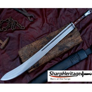Pode incluir: Uma espada longa de gume único com lâmina de aço polido, cabo de madeira escura com rebites prateados e pomo prateado. A espada é acompanhada por uma bainha de couro preto. O texto SharpHeritage Born at the Forge é visível.