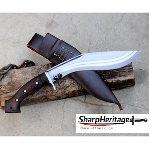 May include: A large, curved blade knife with a dark wooden handle and a leather sheath. The blade is silver and the handle has a cross-stitched design. The sheath is brown and the knife is resting on a piece of wood. The text "SharpHeritage Born at the Forge" is visible.